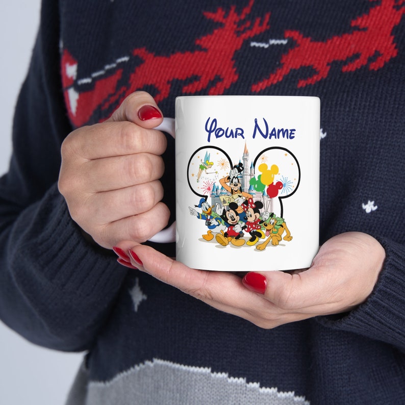 Puede incluir: Taza de cer&aacute;mica blanca con un dise&ntilde;o de personajes de Disney. El dise&ntilde;o incluye a Mickey Mouse, el Pato Donald y otros personajes, con el texto "Your Name" en azul. La taza est&aacute; sostenida por una persona.