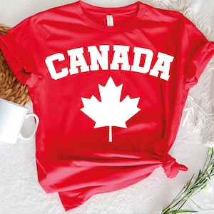 May include: A red t-shirt featuring the word "CANADA" in bold white letters above a white maple leaf graphic. The shirt is made of a soft material and has short sleeves.