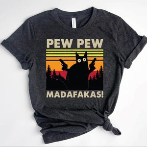 May include: Dark gray t-shirt featuring a black cat silhouette holding two guns, with the text "PEW PEW MADAFAKAS!" in white. The design includes a sunset-themed background with yellow, orange, and black stripes and trees.