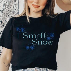 Puede incluir: Camiseta negra con el texto "I Smell Snow" en azul claro, acompañada de gráficos de copos de nieve azules. La camiseta es de manga corta y cuello redondo.