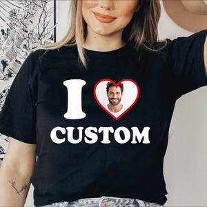 Peut inclure: T-shirt noir avec l'inscription "I CUSTOM". La lettre "I" est blanche, et le mot "CUSTOM" est également blanc. Un cœur rouge avec un bord blanc contient la photo d'une personne souriante.