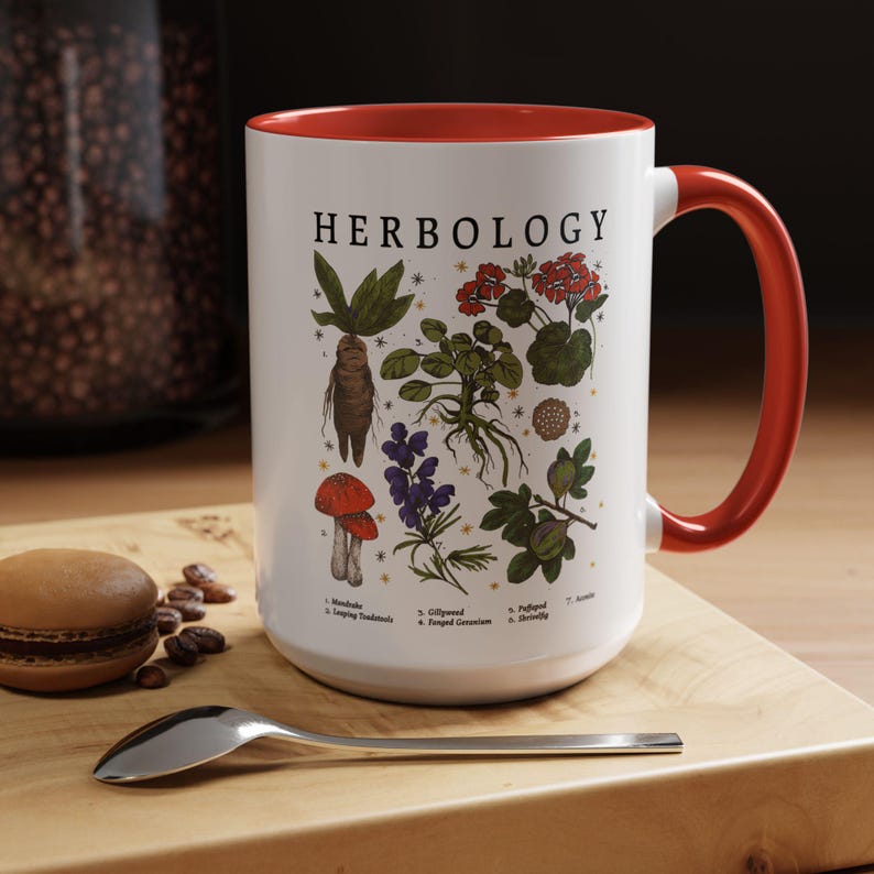 A white mug with a red handle, featuring illustrations of various plants and flowers, is placed on a wooden surface next to a spoon and some coffee beans.