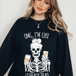 Op de afbeelding: Een zwart sweatshirt met lange mouwen en een witte afbeelding van een skelet met een rommelige knot. Het skelet houdt twee bekers vast, één met een oranje pompoenontwerp en één met oranje tekst. De tekst luidt: OMG, I'M LIKE LITERALLY DEAD.