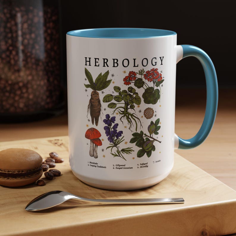 A white mug with a blue handle, featuring illustrations of various plants and flowers, placed on a wooden surface next to a spoon and some coffee beans.