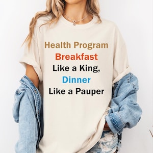 May include: A beige short-sleeved t-shirt featuring the text Health Program in brown, Breakfast in orange, Like a King, in black, Dinner in blue, and Like a Pauper in black. The shirt is worn with a light blue denim jacket and denim shorts.