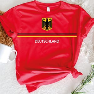 May include: A red t-shirt with the German coat of arms and the word "DEUTSCHLAND" in white. A black and yellow stripe is across the chest. A white mug is visible in the background.