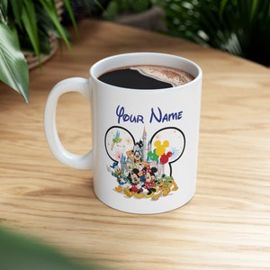 Puede incluir: Taza de cer&aacute;mica blanca con asa negra, llena de caf&eacute;. La taza presenta personajes de Disney como Mickey y Minnie Mouse, Donald Duck y Goofy. Las palabras "Your Name" est&aacute;n escritas encima de los personajes de Disney.