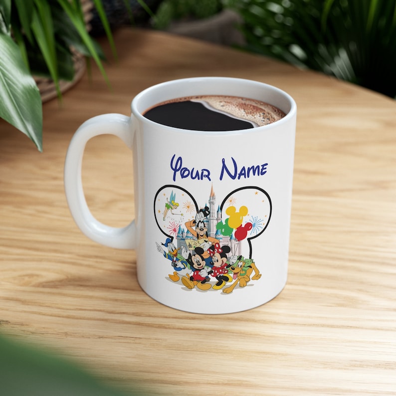 Puede incluir: Taza de cer&aacute;mica blanca con asa negra, llena de caf&eacute;. La taza presenta personajes de Disney como Mickey y Minnie Mouse, Donald Duck y Goofy. Las palabras "Your Name" est&aacute;n escritas encima de los personajes de Disney.