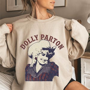 Può includere: Una felpa girocollo beige con una stampa grafica dall'effetto vissuto di Dolly Parton che indossa un cappello da cowboy. Il testo DOLLY PARTON è arcuato in lettere rosso scuro sopra il ritratto, realizzato in tonalità di blu e rosso scuro.