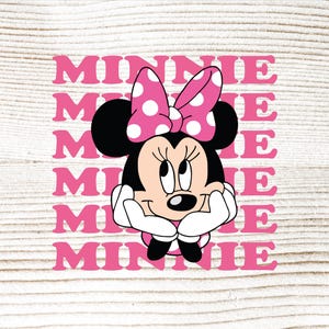 以下が含まれることがあります： ピンクのドット柄のリボンと「MINNIE」の文字がピンク色で繰り返されたミニーマウスのグラフィック。ミニーマウスは黒、白、ピンクで、あごの下に手を置いています。背景は明るい木目です。