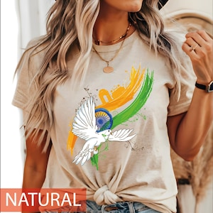 May include: A natural-colored t-shirt featuring a graphic print of a white dove in flight, a blue Ashoka Chakra wheel, and orange and green brushstroke accents. The design includes a silhouette of the India Gate monument and delicate vine details.