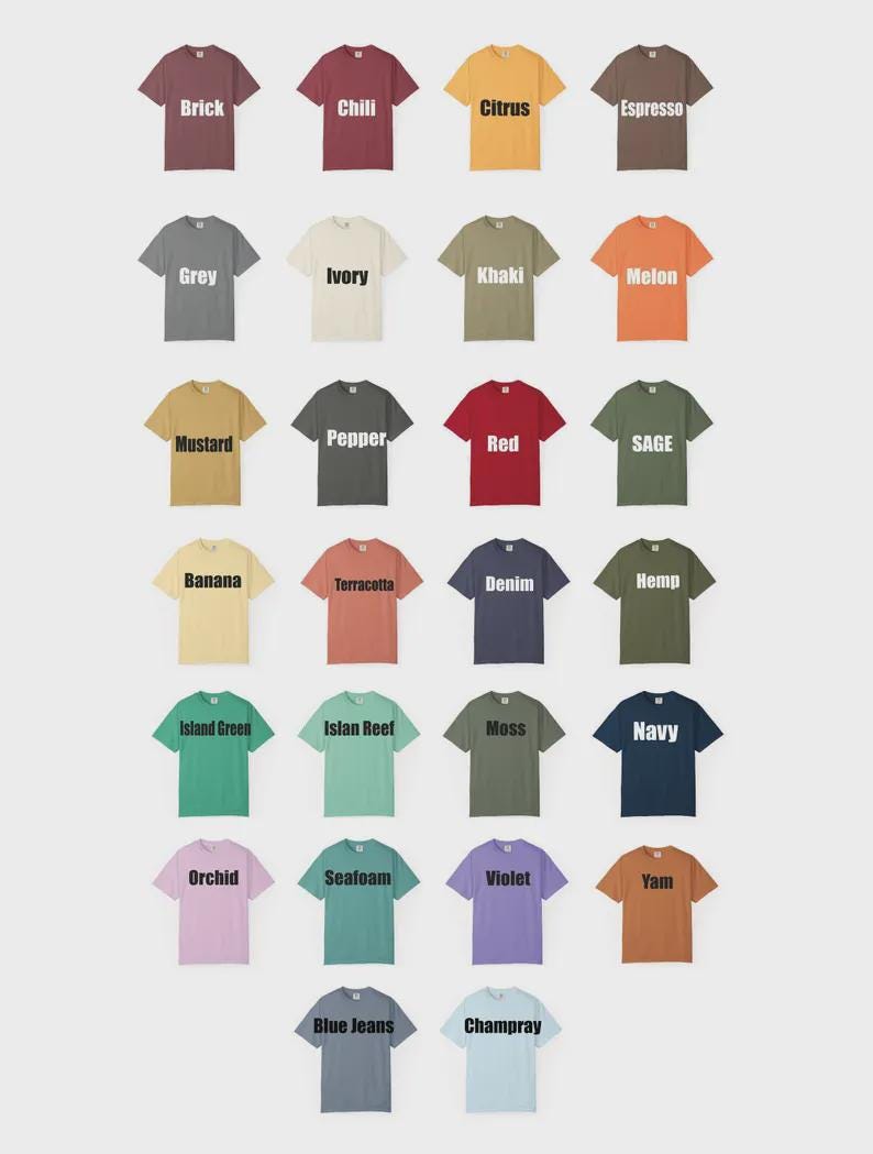 May include: A collection of twenty-six short-sleeve crew neck t-shirts arranged in a grid. Each shirt is a different solid color with the corresponding color name printed in white or black text across the center of the chest area.