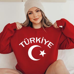 May include: A red sweatshirt featuring the word "TÜRKIYE" in white, accompanied by a white crescent moon and star emblem. The sweatshirt is paired with a beige beanie.