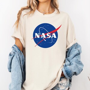 A woman wearing a white t-shirt with the NASA logo and a denim jacket, standing against a white background.