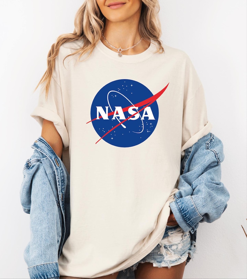 A woman wearing a white t-shirt with the NASA logo and a denim jacket, standing against a white background.