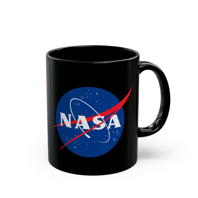 May include: Black ceramic coffee mug featuring the NASA logo. The logo includes a blue circle with white stars and a white text that reads "NASA", with a red swoosh. The mug has a black handle.