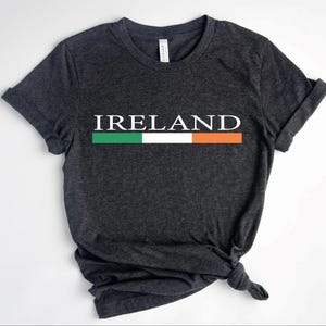 Puede incluir: Camiseta gris oscuro con la palabra "IRELAND" en letras mayúsculas blancas. Debajo, una franja horizontal con los colores de la bandera irlandesa: verde, blanco y naranja. La camiseta está anudada en la parte inferior.