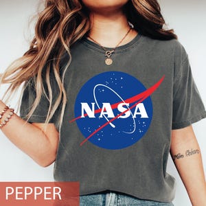 May include: A charcoal gray short-sleeve t-shirt featuring the blue, white, and red NASA logo printed on the center chest. The circular logo includes white text, a white orbital path, and a red vector swoosh against a dark blue background.