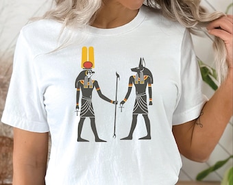 May include: White t-shirt featuring a graphic of two Egyptian deities, one with a falcon head and the other with a jackal head, holding hands. The figures are in black, gray, and gold, with a staff between them. The shirt is a classic crew neck.