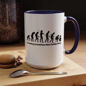Puede incluir: Taza de cerámica blanca con interior y asa azul marino. La taza presenta un gráfico negro que representa la evolución humana, que termina con una persona frente a una computadora. El texto "Something Somewhere Went Terribly Wrong" está debajo.