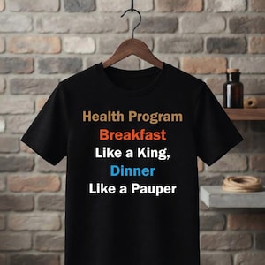 May include: A black short-sleeved t-shirt featuring the text Health Program in tan, Breakfast in orange, Like a King, in white, Dinner in blue, and Like a Pauper in white. The shirt is displayed on a wooden hanger against a brick wall background.