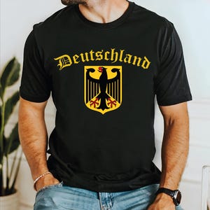 May include: Black t-shirt featuring the word "Deutschland" in gold gothic lettering above a gold and black coat of arms with a black eagle. The eagle has a red beak and feet. The shirt is worn by a person.