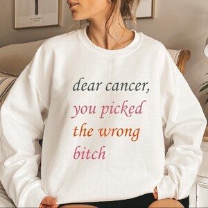May include: A white crewneck sweatshirt featuring the text "dear cancer, you picked the wrong bitch" printed in a multi-coloured font. The word "dear cancer," is in dark grey, "you picked" is in pink, "the wrong" is in orange, and "bitch" is in pink.