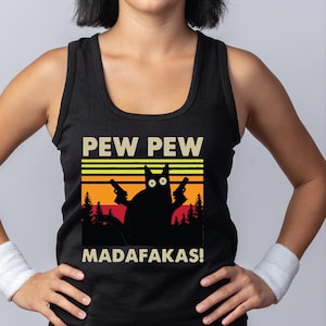 Op de afbeelding: Een zwart hemdje met een grafische print van een zwarte kat die twee handvuurwapens vasthoudt. Het ontwerp bevat een retro zonsondergangachtergrond met silhouetten van dennenbomen en de tekst PEW PEW MADAFAKAS! in een vetgedrukt, licht lettertype.