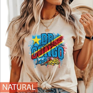 May include: A beige short-sleeve t-shirt featuring a graphic print with the text DR CONGO LEOPARDS. The design includes a yellow star, a leopard silhouette, and blue, red, and yellow colours arranged in a paint-splatter style with drips.