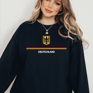 May include: Black sweatshirt with the word "Deutschland" and the colours of the German flag. The design includes a yellow and black eagle crest above a horizontal stripe with red, yellow, and black colours.