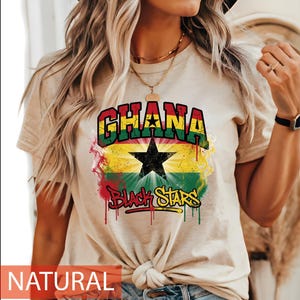 May include: A natural-coloured t-shirt featuring a graphic print of the Ghana flag with a central black star. The text "GHANA" appears in bold letters above the flag, and "BLACK STARS" is written in a stylised, dripping font below the star.