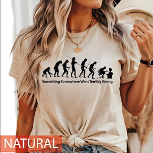 Op de afbeelding: Een naturelkleurig T-shirt met korte mouwen en een zwarte afbeelding van de menselijke evolutie die eindigt met een figuur achter een computerbureau. Onder de afbeelding staat de tekst Something Somewhere Went Terribly Wrong in zwarte letters.