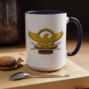 May include: White ceramic mug with a navy blue handle and interior. The mug features a gold emblem of an eagle within a wreath, with the letters "S.P.Q.R." below. The mug is on a wooden surface.