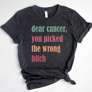 Puede incluir: Camiseta gris oscuro con el texto "dear cancer, you picked the wrong bitch" en verde azulado, rosa y naranja. La camiseta está hecha de un material suave y tiene las mangas dobladas.