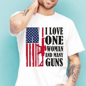 May include: White t-shirt with a graphic design featuring the American flag and a rifle silhouette. The text reads "I LOVE ONE WOMAN AND MANY GUNS" in black. The shirt is worn by a person.