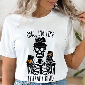 May include: A white short-sleeved t-shirt featuring a black graphic of a skeleton with a messy bun, holding a pumpkin-decorated cup and a cup with the letters AFF. The text reads, OMG, I'M LIKE LITERALLY DEAD, in black capital letters.