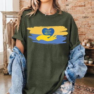 以下が含まれることがあります： オリーブグリーンのTシャツ。黄色と青のブラシストロークの背景に、黄色い手が青いハートを支えているグラフィックが描かれています。ハートの中には黄色い文字で「I STAND WITH UKRAINE」とプリントされています。