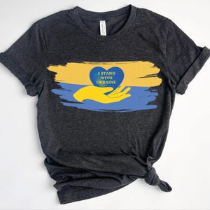 以下が含まれることがあります： ダークグレーの半袖Tシャツ。黄色い手が青いハートを持っているグラフィックが描かれています。ハートの中には黄色い文字で「I STAND WITH UKRAINE」と書かれています。グラフィックは黄色と青の筆跡の背景の上に配置されています。