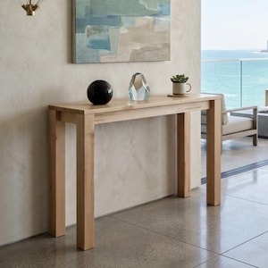 May include: A light-toned wooden console table with four straight legs and a rectangular top. On the surface sits a smooth black sphere, a clear geometric glass sculpture, and a small green succulent plant in a white ceramic mug.