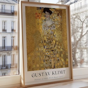 Puede incluir: Una impresión enmarcada del "Retrato de Adele Bloch-Bauer I (1907)" de Gustav Klimt descansa sobre un alféizar de madera. La obra de arte presenta a una mujer con un vestido dorado con intrincados patrones. El marco es de color marrón claro, con el texto "GUSTAV KLIMT".