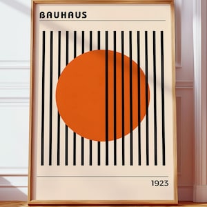 Op de afbeelding: Een ingelijste Bauhaus-kunstprint. Het kunstwerk toont een grote oranje cirkel, zwarte verticale lijnen en het woord "BAUHAUS" bovenaan. Het jaar "1923" staat onderaan afgedrukt. De lijst is lichtbruin.