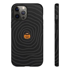 May include: A black smartphone case featuring a dark gray concentric circular wave pattern across the back and sides. Centered on the back is a small orange jack-o'-lantern wearing a black witch hat with a gold buckle detail.
