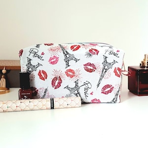 Peut inclure: Une trousse de toilette blanche avec un imprimé tour Eiffel noir et blanc et des lèvres rouges. Le sac porte l'inscription "Paris" plusieurs fois.