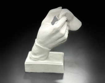 May include: A white plaster sculpture of a hand holding a small, rounded object. The hand is detailed, with visible fingers and a sleeve. The sculpture is mounted on a rectangular base, set against a dark background.