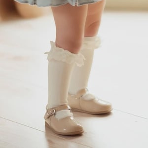 May include: Cream-colored knee-high socks with a delicate lace trim are paired with shiny, light brown Mary Jane shoes. The shoes have a buckle closure and a rounded toe. The image shows a child's legs and feet.