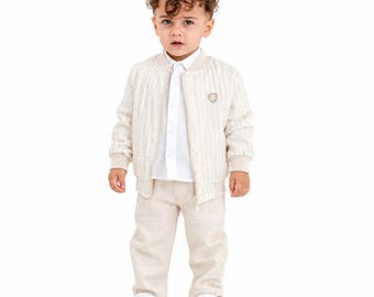 May include: A beige striped bomber jacket with ribbed cuffs and hem, worn over a white button-down shirt and matching beige trousers. The outfit is completed with white sneakers, featuring a small emblem on the left chest of the jacket.