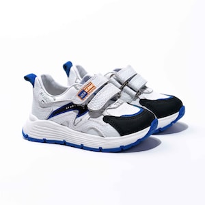 May include: White leather sneakers with blue and black accents. The shoes have two velcro straps and the text "SPORT SHOES" is printed on the side.