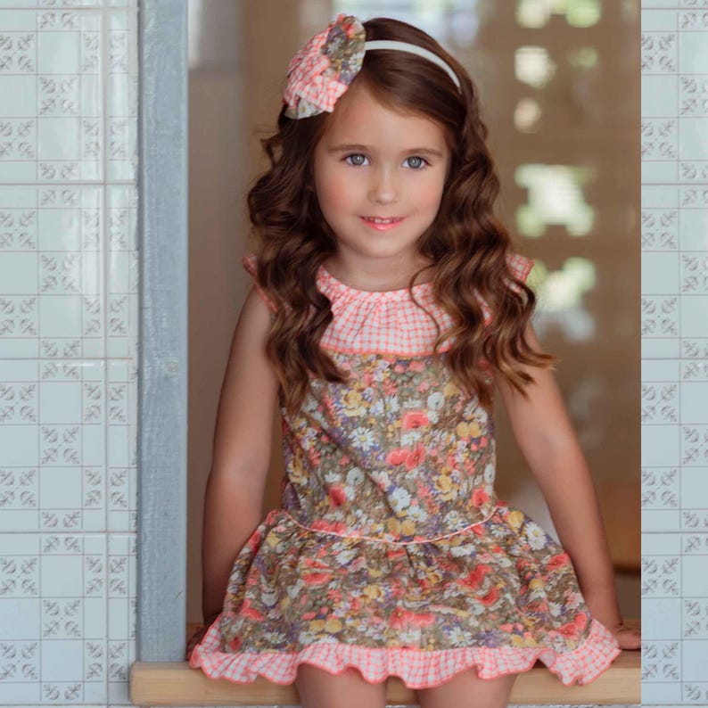 May include: A young girl in a floral dress with a pink gingham collar and hem. She wears a matching floral headband with a white band. The dress has a brown base with colourful flowers, creating a charming, vintage look.