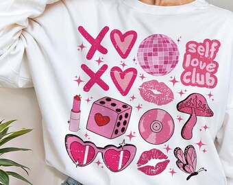 May include: White sweatshirt with a pink graphic design. The design includes hearts, lips, a disco ball, a mushroom, a butterfly, and the words "self love club". The sweatshirt is a casual, comfortable style.