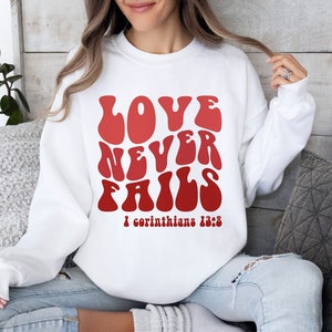 May include: White sweatshirt with a red graphic that says "LOVE NEVER FAILS" in a retro font. The graphic also includes the text "1 Corinthians 13:8".